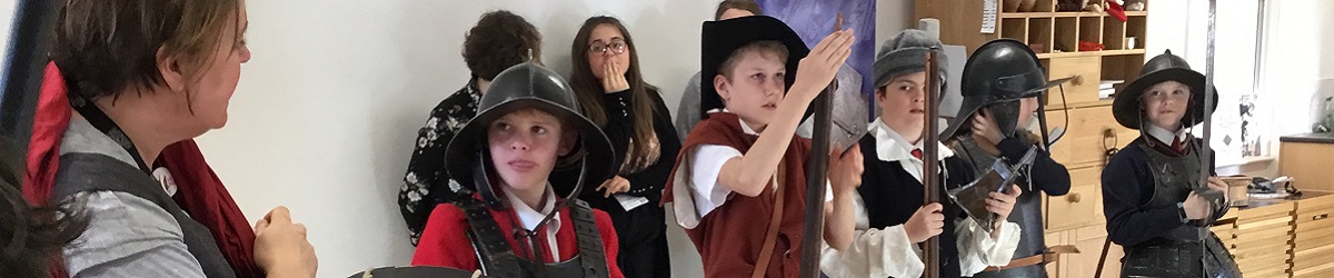 Five young students stand in a line, dressed in Civil War armour and accessories, listening to a member of the National Civil War Centre Learning Team as she teaches them how to use their replica weapons